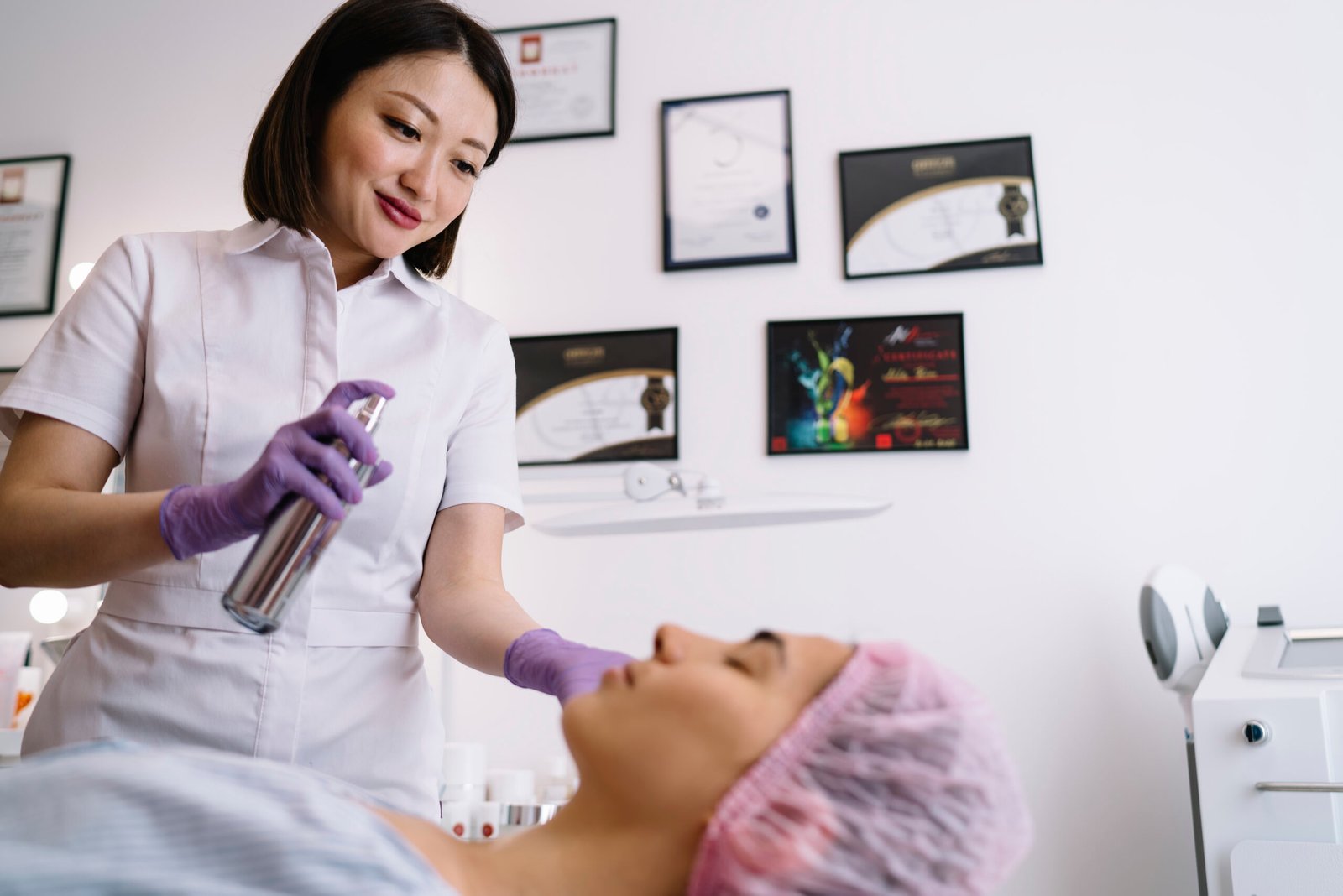 Positive Asian female beautician spraying water on client Low angle of content young Asian female cosmetician in protective gloves smiling and spraying thermal water on face of client after beauty procedure