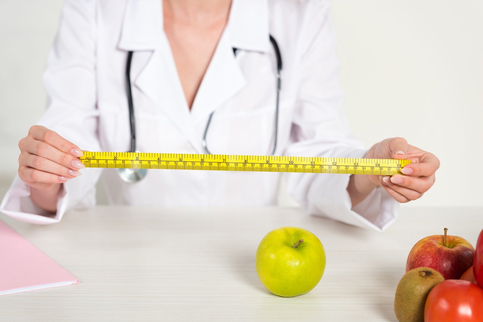 cropped view of dietitian in white coat holding measure tape and fresh food on table