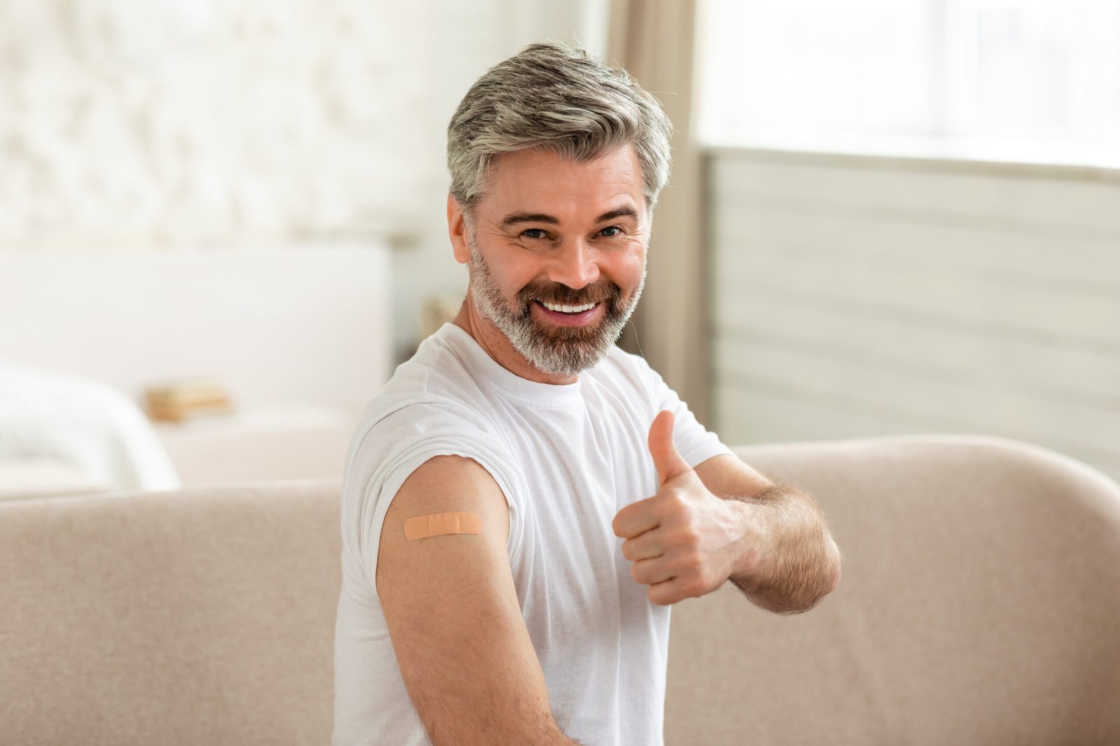 Coronavirus Protection. Cheerful Vaccinated Male Showing Medical Adhesive Plaster Bandage On Arm And Gesturing Thumbs Up Smiling To Camera Sitting Indoor. Covid-19 Vaccination Concept