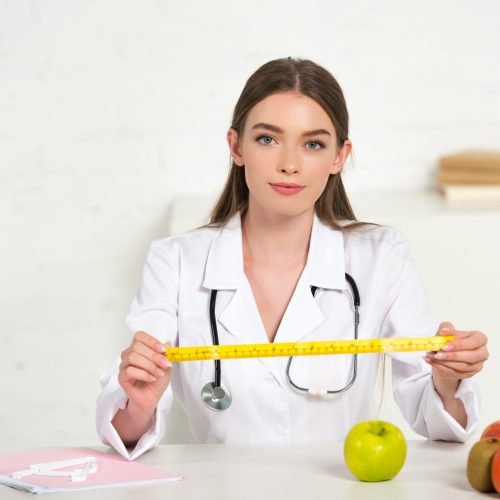 front view of dietitian in white coat with stethoscope holding measure tape at table with fruits and