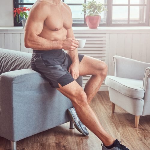 Handsome shirtless muscular male lying on the sofa, drink cup of tea, resting after a hard workout at home.