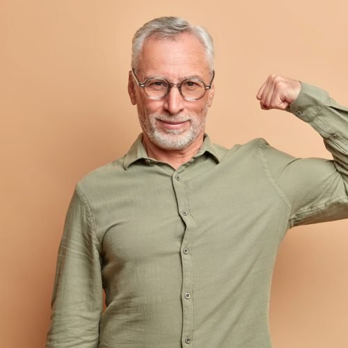 Self confident serious man raises arm shows muscles being confident in his strength wears formal shirt poses against brown background. Healthy handsome European pensioner demonstrates biceps