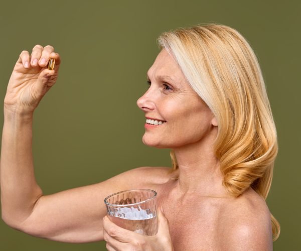 A smiling mature woman holds a supplement and a glass of water, advocating for healthy lifestyle choices and wellness. She exudes positivity and vitality