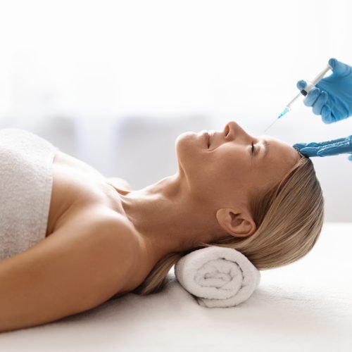 Injection Cosmetology. Beautician Doctor Making Beauty Shot To Forehead Of Middle Aged Woman, Attractive Mature Female Getting Interbrow Anti-Wrinke Treatment While Lying On Table In Spa Salon