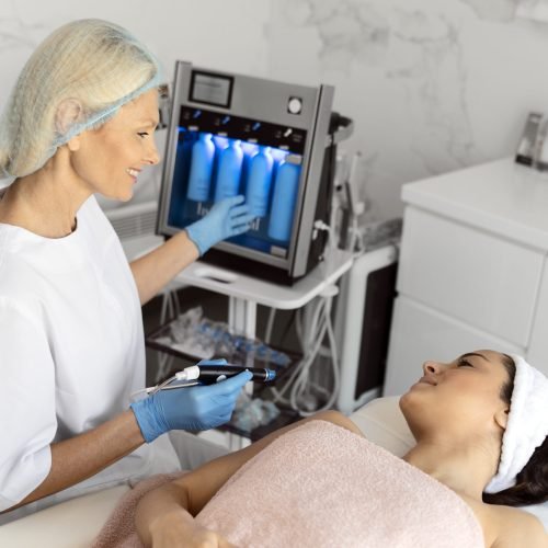 Cleansing procedure. Nice pretty woman lying with her eyes closed while having a cleansing procedure on her face. Senior doctor pointing on the screen while working with smile