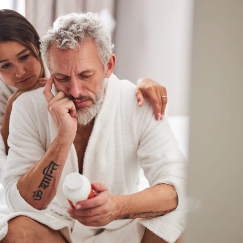 Close up front view portrait of bearded Caucasian male in white bathrobe looking to the pill jar while sitting on the bedroom with his wife