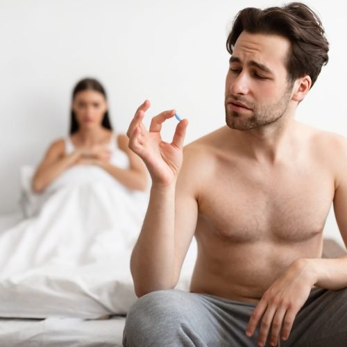 Impotence Issue. Young Husband Holding Potency Pill Before Sex While Wife Waiting In Bed Indoors. Medicine For Male Sexual Health Problems. Low Libido Treatment Concept. Selective Focus
