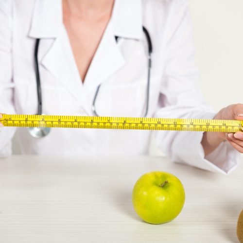 cropped view of dietitian in white coat holding measure tape and fresh food on table