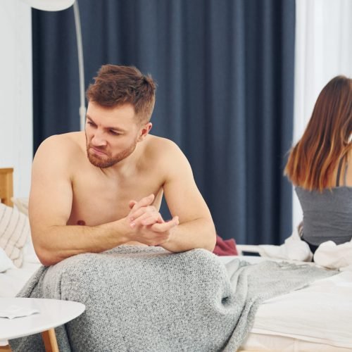 Disappointed woman sitting on the bed with back to the man that feels bad.