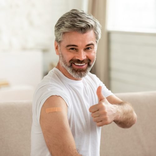 Coronavirus Protection. Cheerful Vaccinated Male Showing Medical Adhesive Plaster Bandage On Arm And Gesturing Thumbs Up Smiling To Camera Sitting Indoor. Covid-19 Vaccination Concept