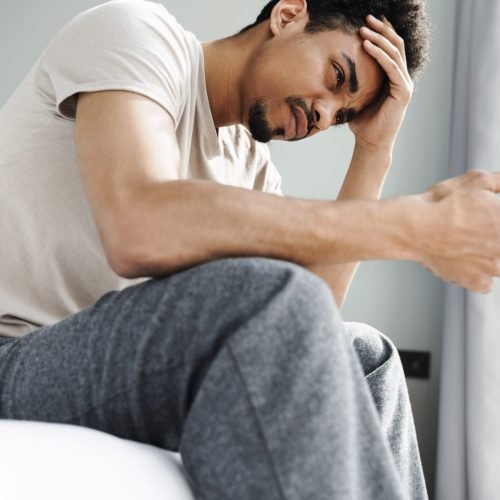 Photo of displeased african american man using mobile phone while sitting on bed after sleep at bright room