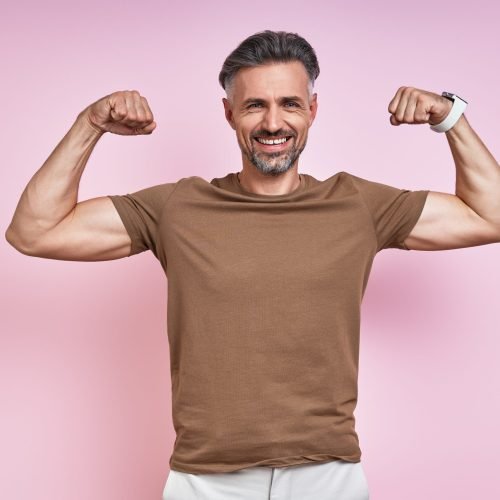Happy mature man showing his biceps while standing against pink background